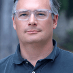 Dr. Jeffrey Lewis, man with grey and black hair wearing clear plastic frame glasses and a blue collard shirt, looks into camera.