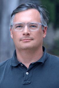 Dr. Jeffrey Lewis, man with grey and black hair wearing clear plastic frame glasses and a blue collard shirt, looks into camera.