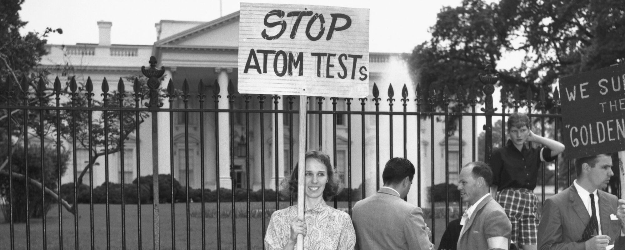 A woman demonstrating outside the White House holds a picket sign protesting nuclear testing, while several men gather nearby. The woman's sign reads "STOP ATOM TESTS."